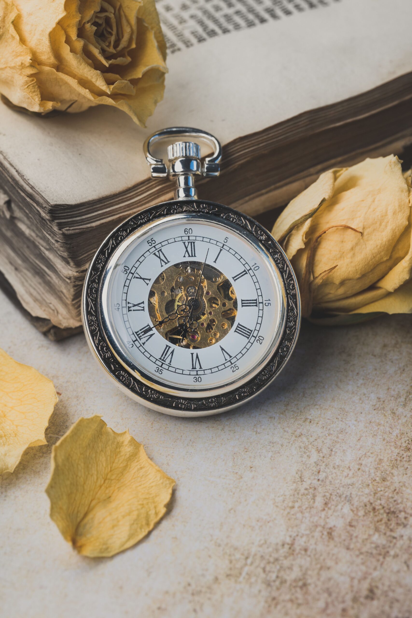 A vintage pocket watch rests on a book with dried yellow roses, creating an antique ambiance.